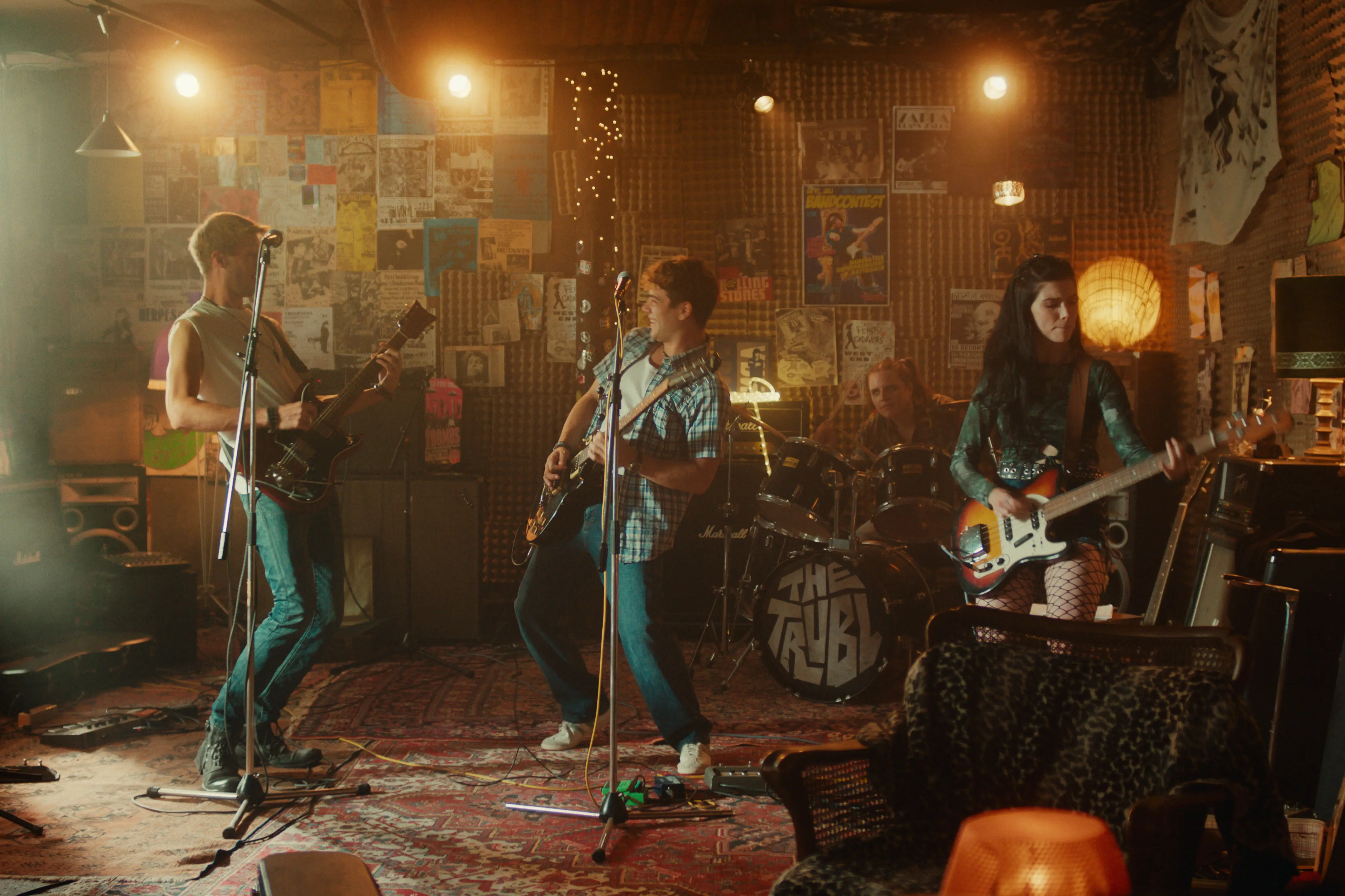 Vier junge Musiker spielen Gitarre und Schlagzeug in einem schwach beleuchteten, gemütlichen, mit Postern bedeckten Proberaum.
