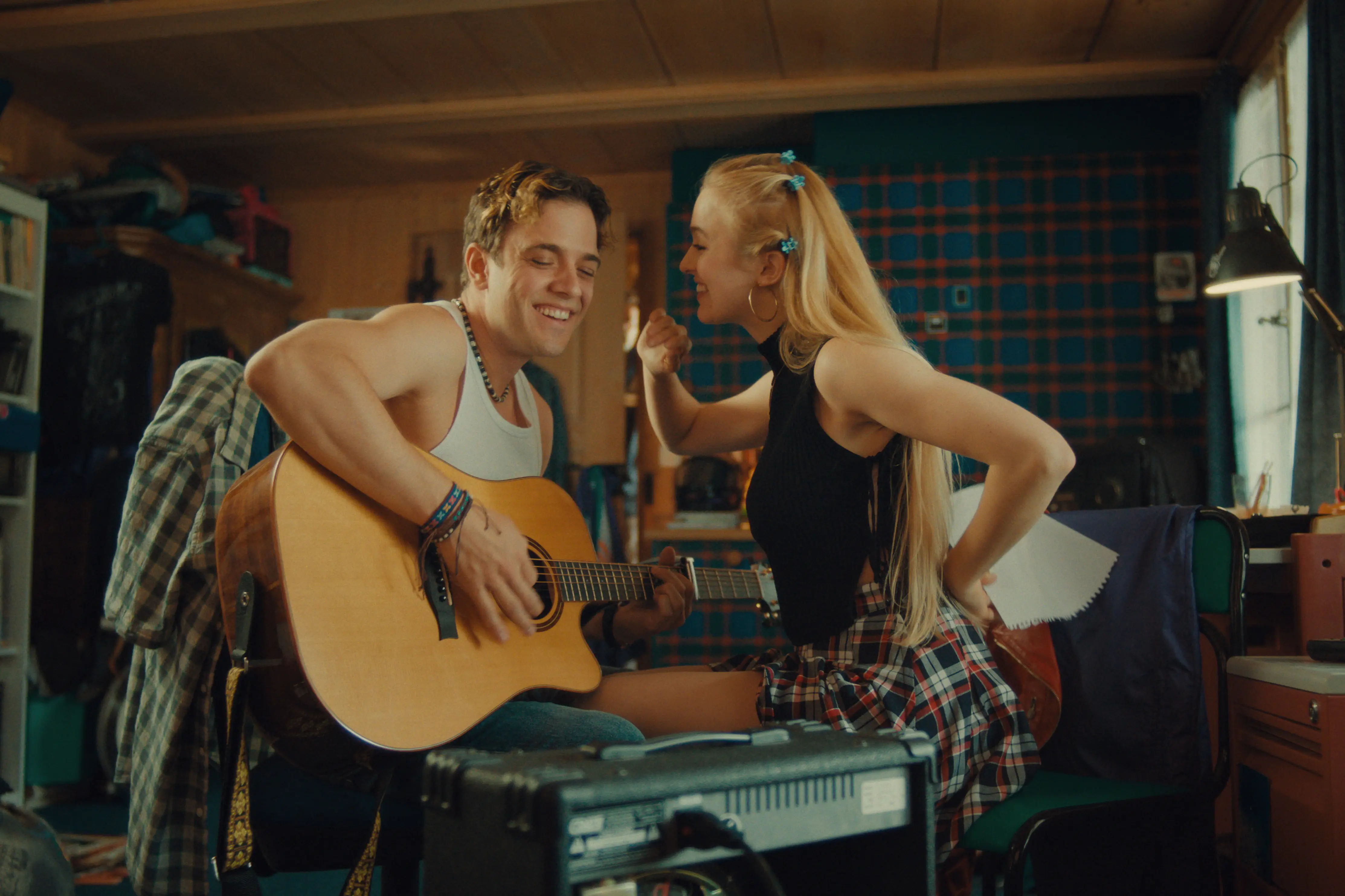 Ein junger Mann spielt Gitarre und lächelt, während neben ihm in einem gemütlichen Zimmer eine junge Frau mit langen blonden Haaren und kariertem Rock fröhlich gestikuliert.