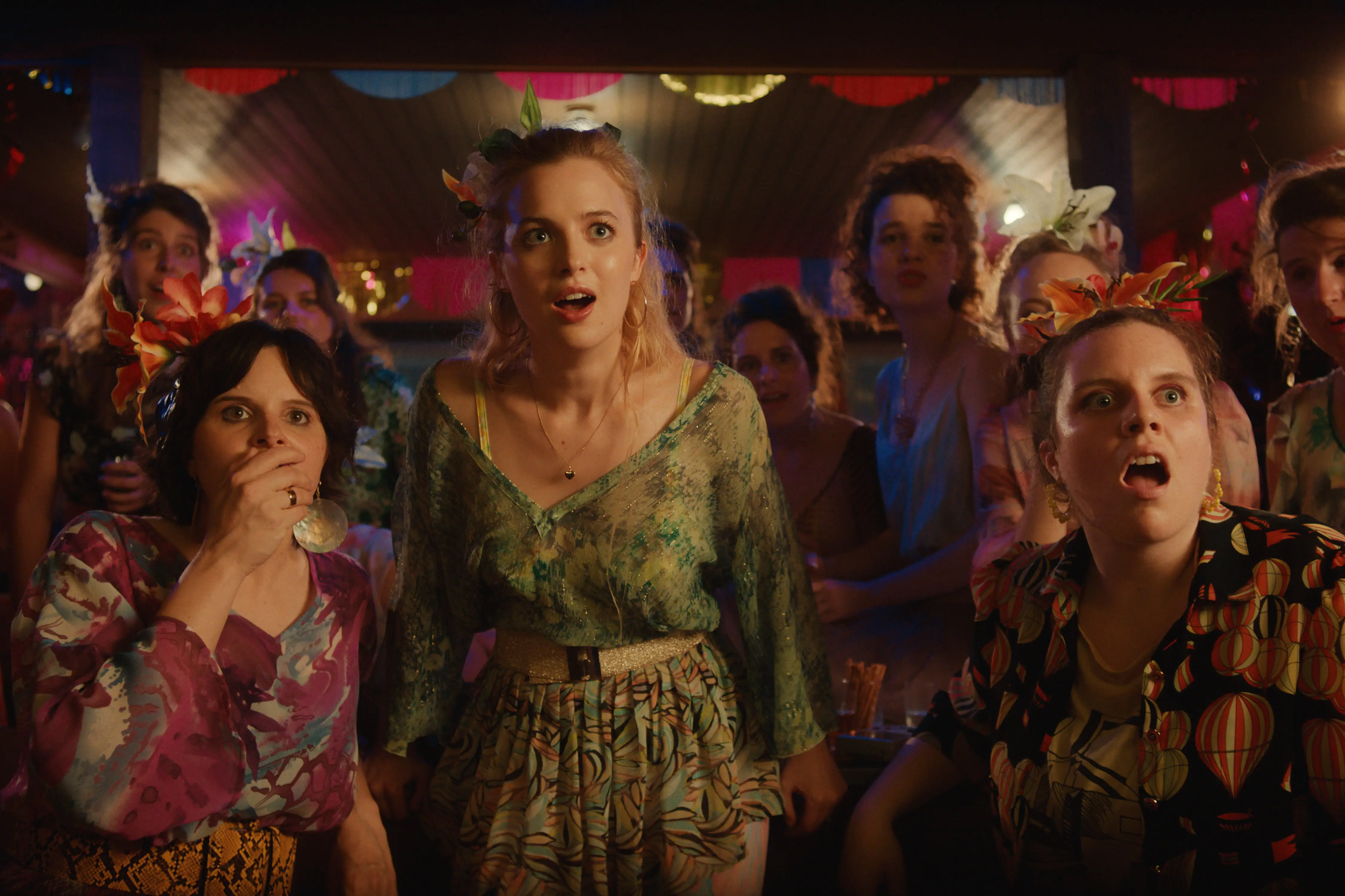 Eine Gruppe von Frauen auf einer Party, in farbenfrohen Blumenkleidern und Kopfbedeckungen, alle mit Gesichtsausdruck der Überraschung und des Staunens.
