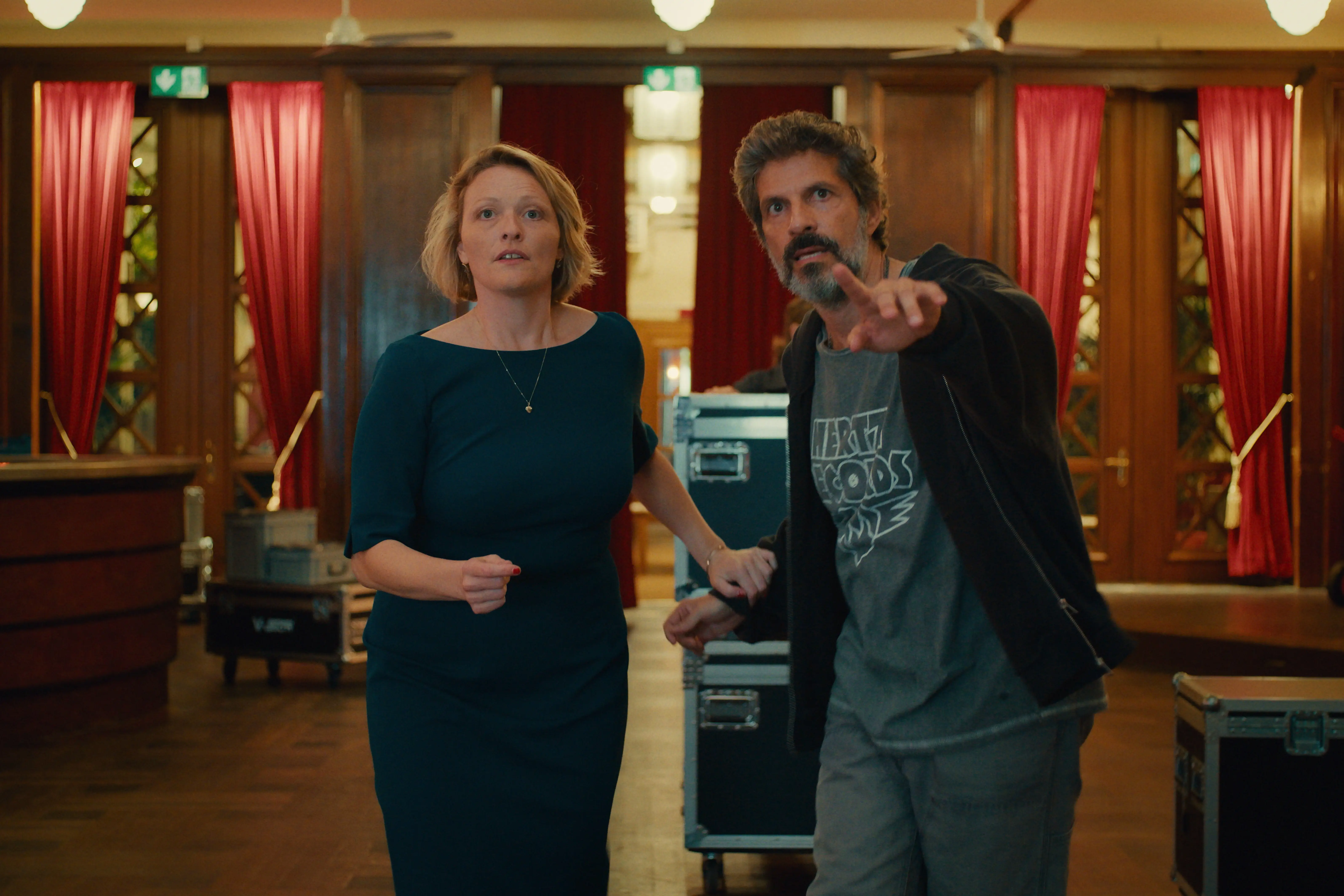 Eine Frau in einem dunkelgrünen Kleid und ein Mann in Freizeitkleidung blicken konzentriert nach vorn, wobei der Mann in einem holzgetäfelten Raum mit roten Vorhängen nach vorn zeigt.
